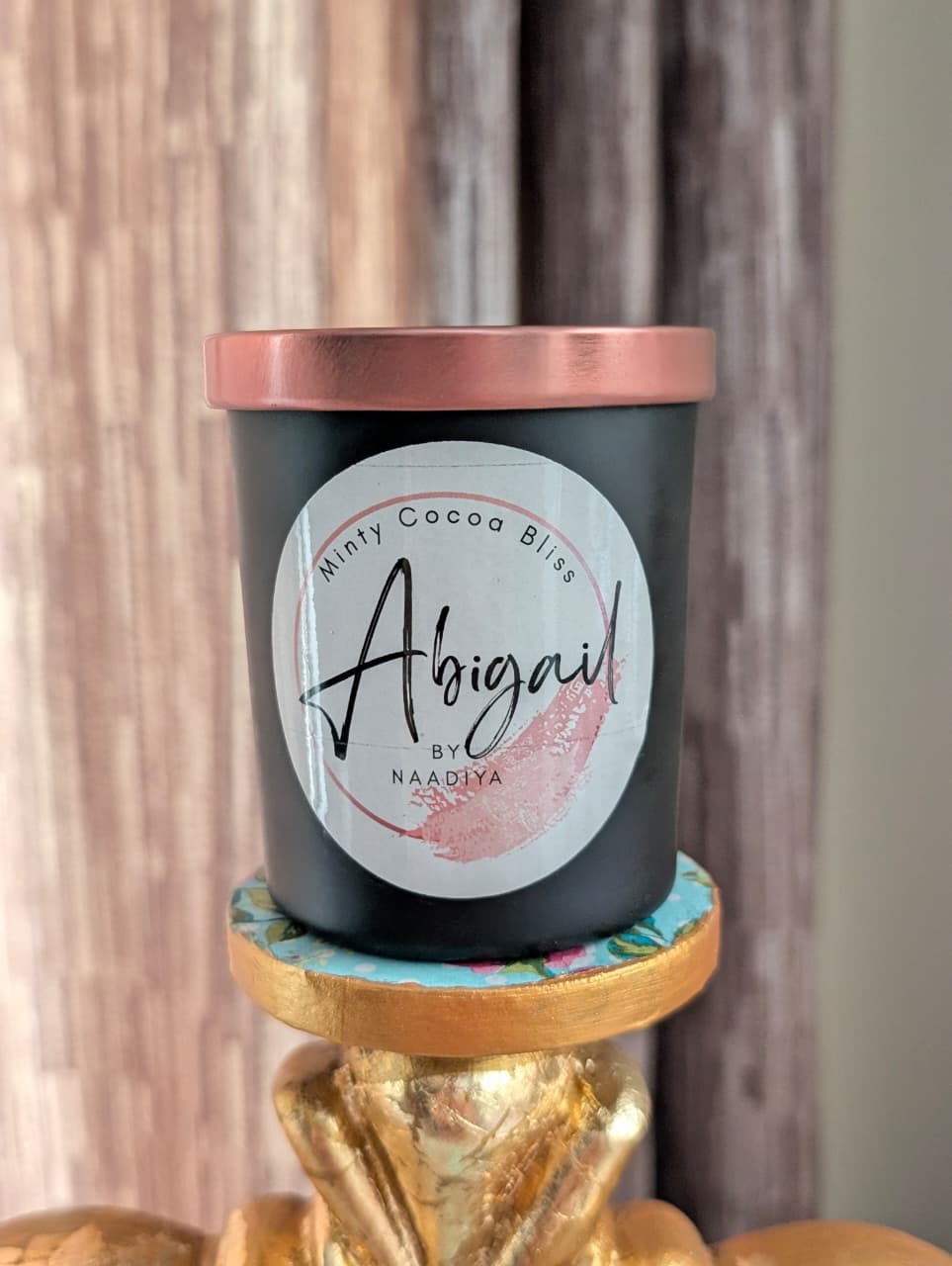 Black candle with a rose gold lid on a decorative stand against a neutral background